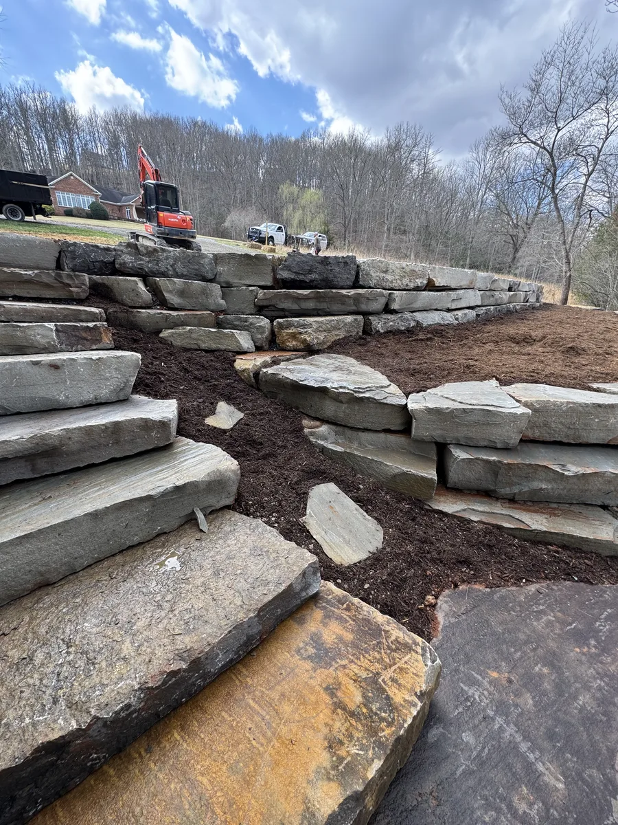 Stacked gray boulders forming a wall beside stone steps and a house
